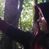 Profile of white non-binary person, facing to the left and holding up their arm against a tree. They are wearing a black hoodie with red trim, the hood pulled up. Some brown hair peeks out from under the hood. Their eyes are shut against the sunlight fi